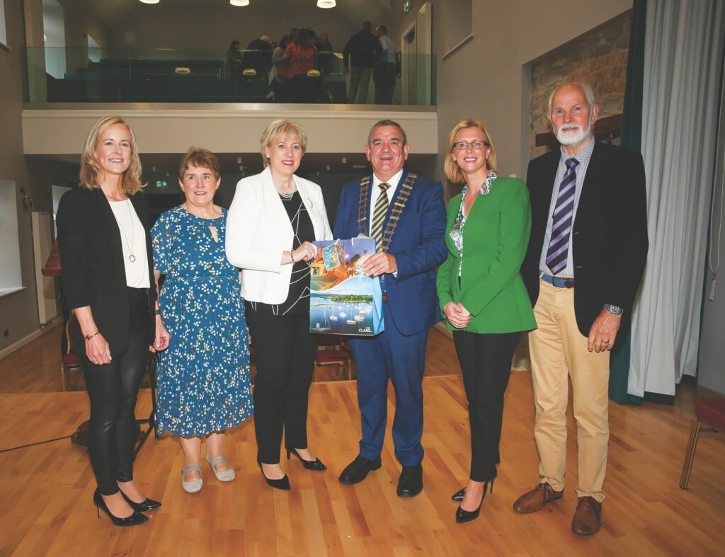 A group of six formally dressed people, in the middle a woman and a man hold a gift bag between them, the man is wearing a golden mayoral chain