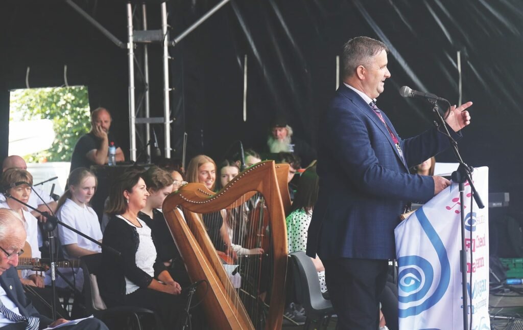 A man stands talking into a microphone on a stand at a podium covered in white fabric with the Comhaltas logo, he is on an outdoor covered stage, behind him sit adults and children, some have instruments including a harp and guitar