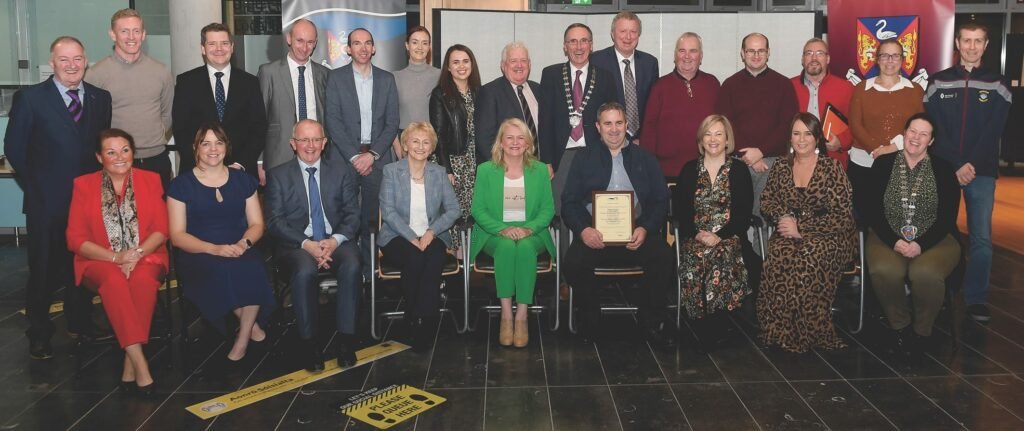 A large group of adults, the front row seated, the others standing behind, some people wear suits or blazers, others dress in a range from very casual to evening wear, a man in the back row wears a golden mayoral chain, a man in the front row holds a framed certificate