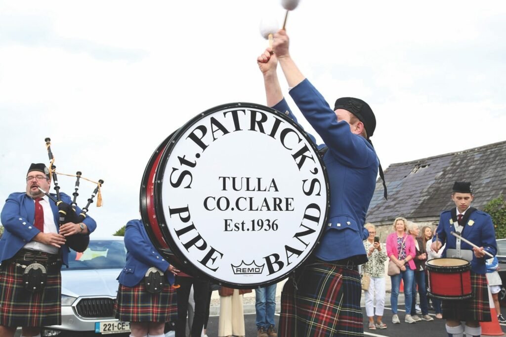 A man stands in the middle with drumsticks held up above a large drum with St Patrick's Pipe Band written on the side, two more musicians on either side are playing bagpipes and a smaller drum, another member is in partial view, they are wearing mainly red and black tartan kilts, a blue jacket, black hats with a silver medallion on the side