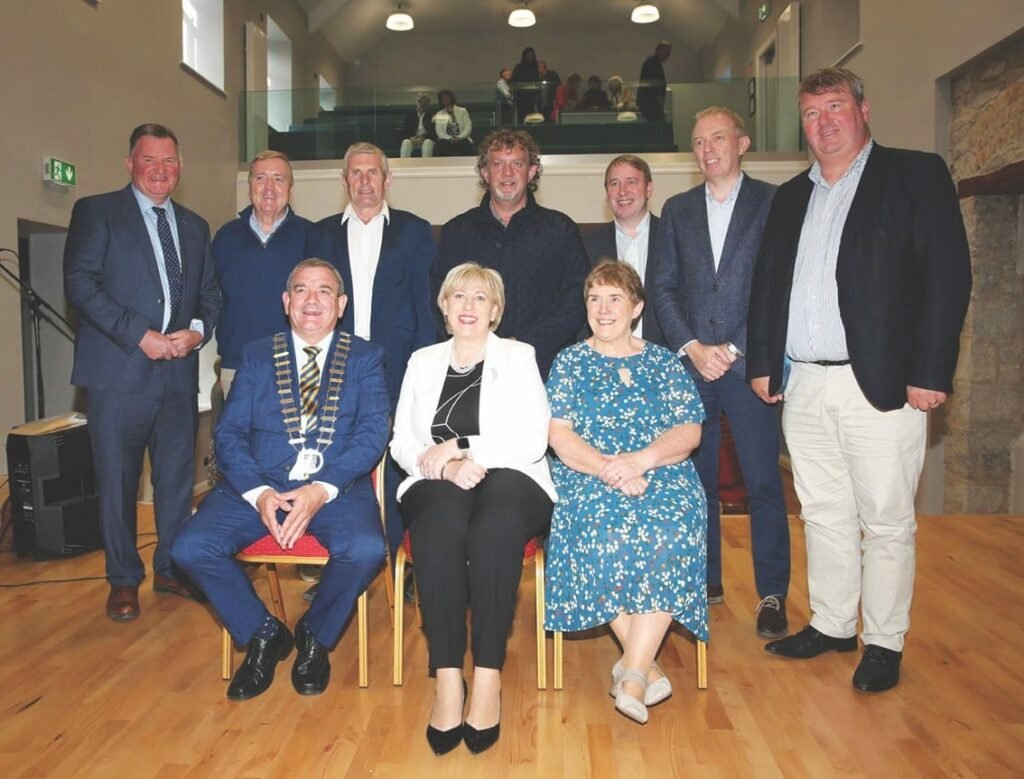 A man in a golden mayoral chain sits beside two women, seven men stand behind, the women and most of the men are formally dressed, the other men are casually dressed.