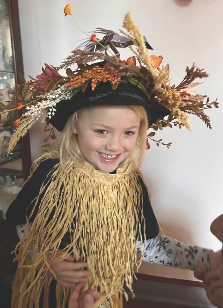 A smiling child wears a hat decorated with grasses and flowers and a bird