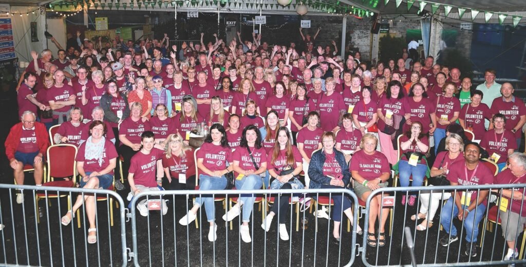 Fleadh Volunteers Mullingar 2022. (Image courtesy of John McCauley) - Many rows of seated people behind temporary event fencing, some smiling, some waving at the back, most are wearing burgundy red t-shirts with printed words on the front