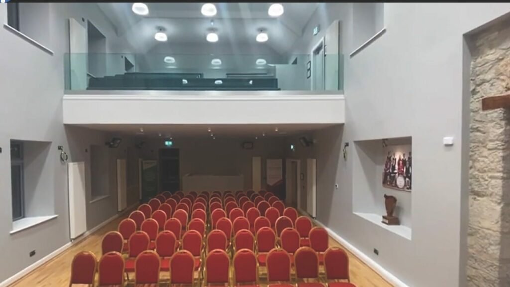 An empty hall with grey walls and a wooden floor, with rows of red chairs, above is a glass-fronted balcony with black seating visible