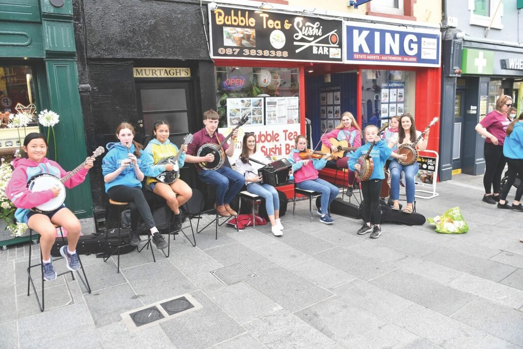 Children and young adults playing music in the street outside shops, all are seated except one of the banjo players is standing, other instruments being played are guitar, accordian and violin