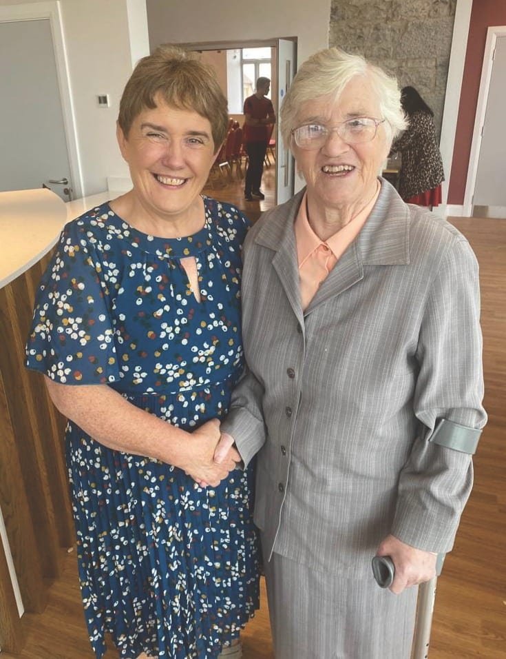 Two smiling women holding or shaking hands, the one on the left wearing a blue patterned dress, the one on the right a grey suit, and she holds a crutch on her left side