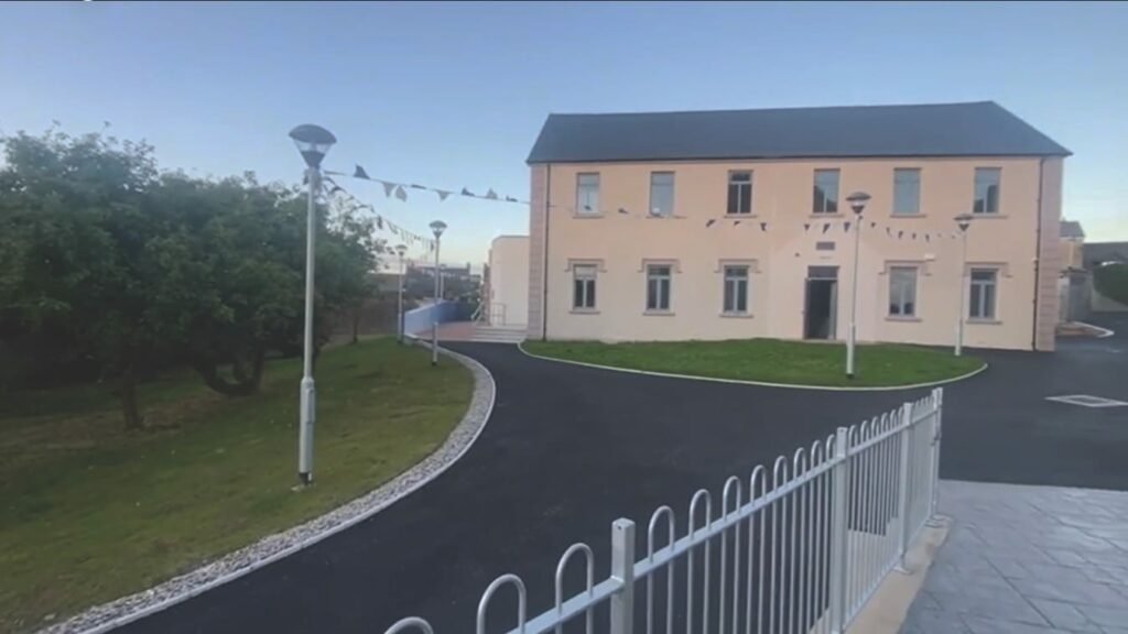 A large beige building, in front are black roads or paths, trees and grass to the left, lamp posts with bunting strung between, and a fence in the foreground of the image.