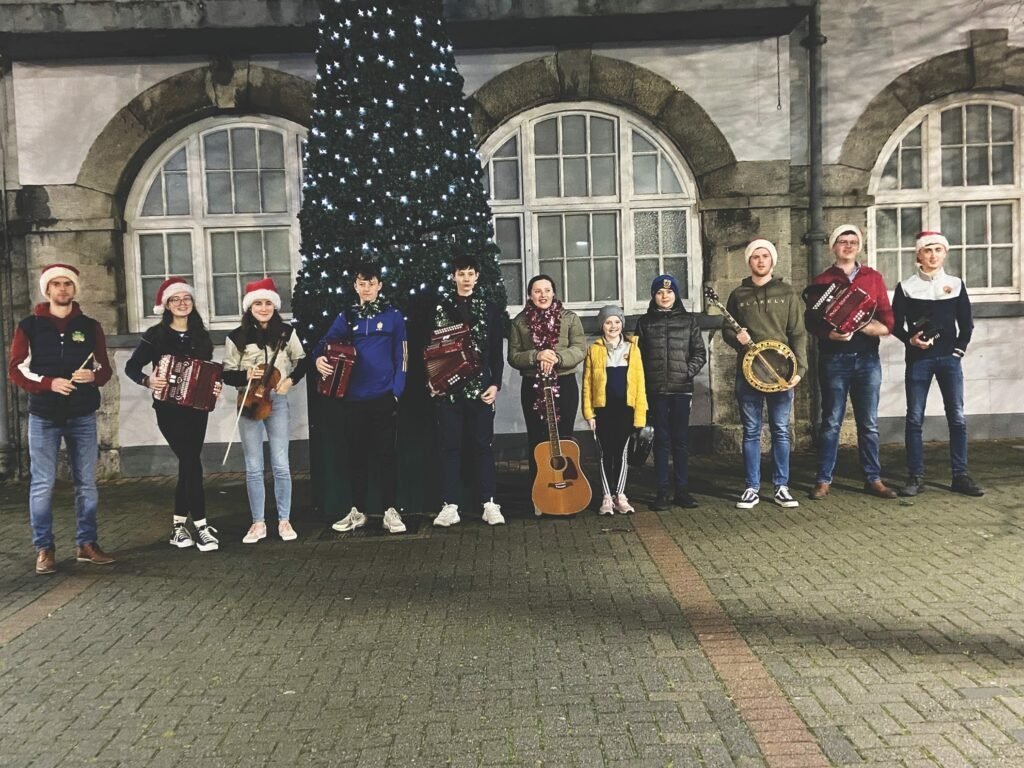 A group of children and young adults are standing, most are holding instrument, they are wearing santa hats and tinsel, behind them is  a large Christmas tree decorated with baubles, and large arched windows of a stone building