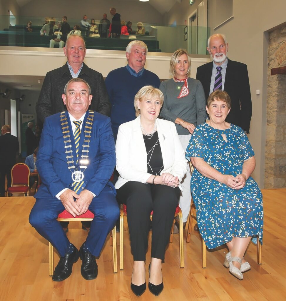 A man in a golden mayoral chain sits beside two women, three men and a woman stand behind, some people are formally dressed, some are casually dressed.