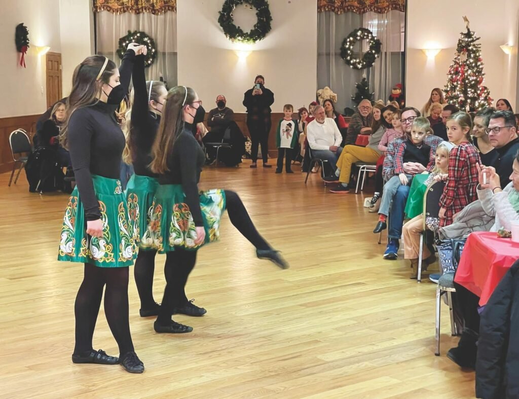 Inside a room with a Christmas tree and decorations Irish dancers dressed in black with green embroidered skirts and performing for an audience