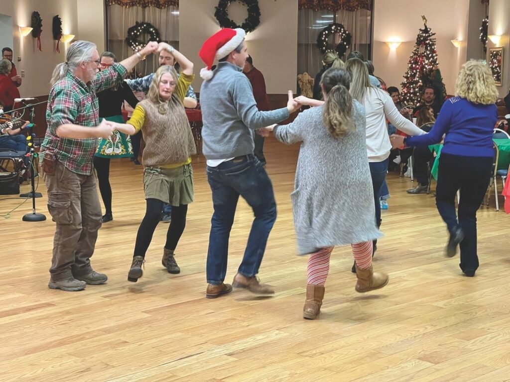 A group of people dancing inside in pairs holding hands, one is wearing a red Santa hat with a white border and pompom