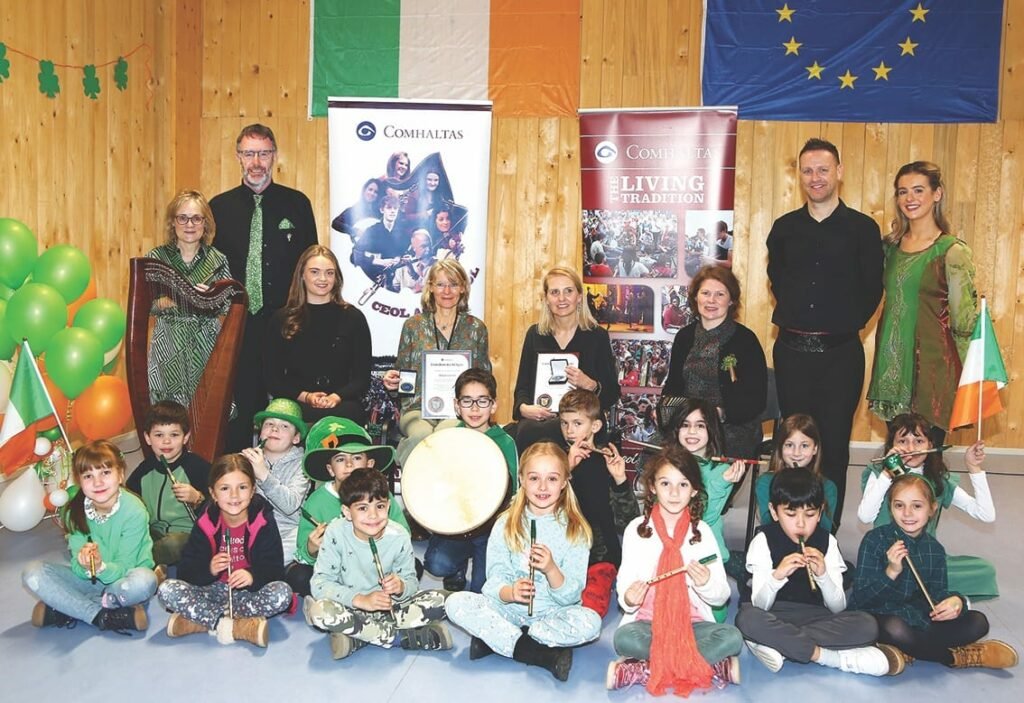 A group photo of children sitting on the floor holding instruments, behind them adults are sitting on chairs or standing, two are holding medals and certificates, there is a harp standing on the floor, an Irish flag is on the wall, another on the floor on the left with green, orange and white balloons, and a child to the right holds an Irish flag, also on the wall behind are an EU flag and two large Comhaltas posters