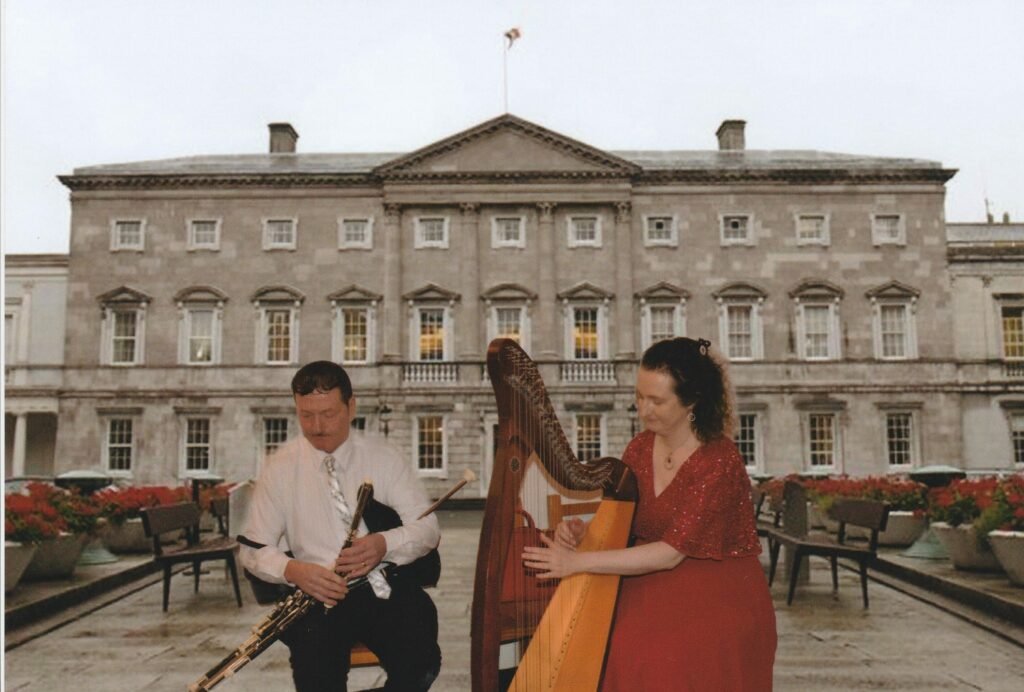 A woman and man sitting outside a large mansion, she is dressed in red and playing harp, he is in a white shirt and tie playing uilleann pipes bagpipes