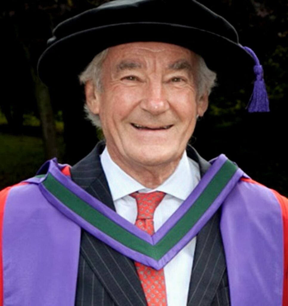 Colour photo of a smiling man in a suit and red tie, wearing a purple, red and green graduation gown and a black hat with a purple tassel hanging to his left