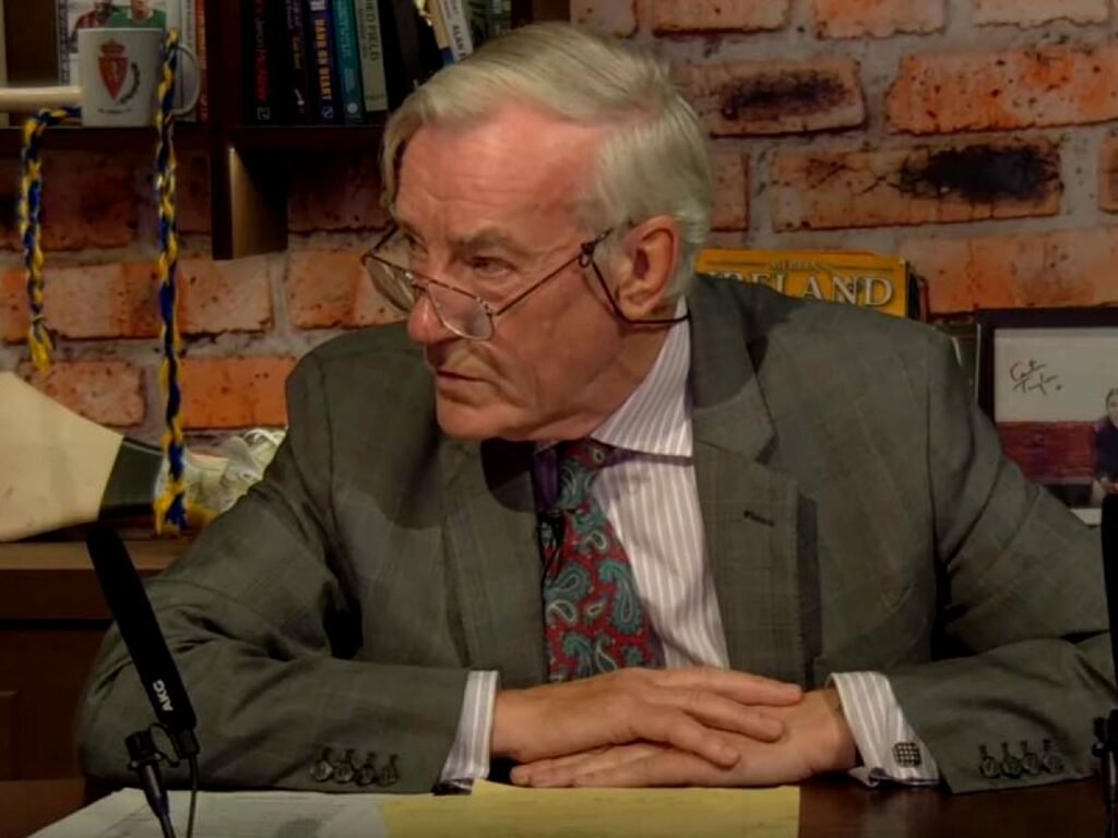 Colour photo of a man in a suit wearing glasses, sitting with his hands on a table in front of him, looking his right, a microphone on the table in front of him