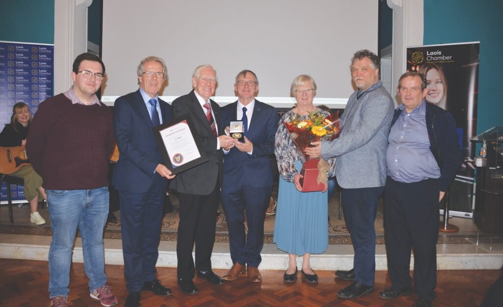 Seven smiling people standing, four men, two holding a medal between them, one holding a framed certificate, are to the left of a woman wearing a blue skirt and patterned top, holding a box with a ribbon bow, two men are to the right, the closest is presenting her with a large bouquet of flowers, in the background to the left sits a woman with a guitar