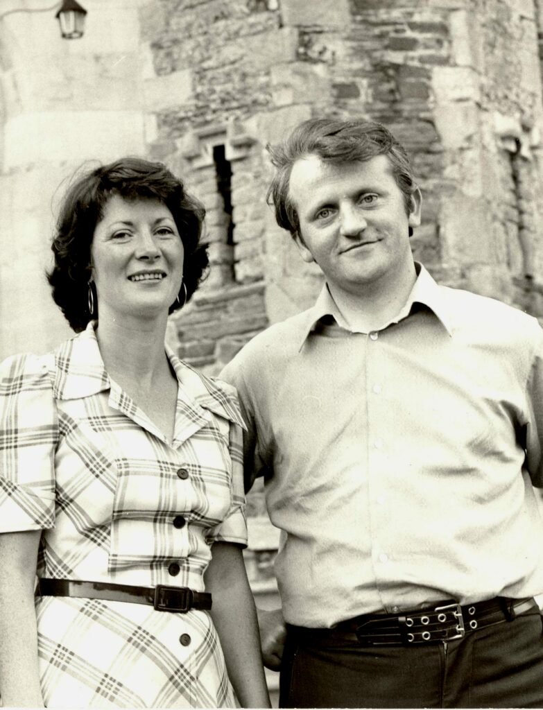 Black and white photo of woman in a check shirt and man in a light shirt, standing in front of an old stone building