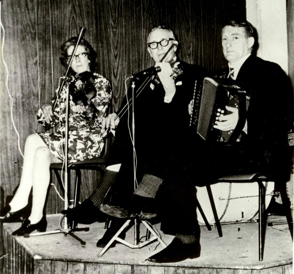 An old black and white photo of three people sitting on a small stage, the woman and a man play violin, another man plays accordian