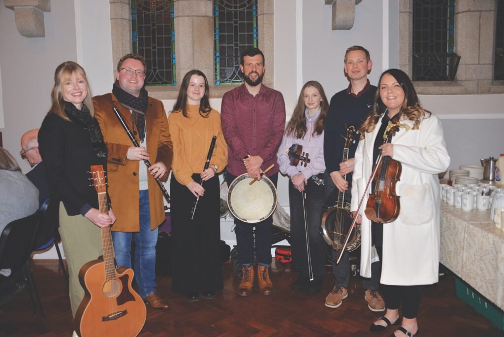 A group of adults and children stand in a building with stone walls and stained glass windows, all holding instruments, some are wearing coats