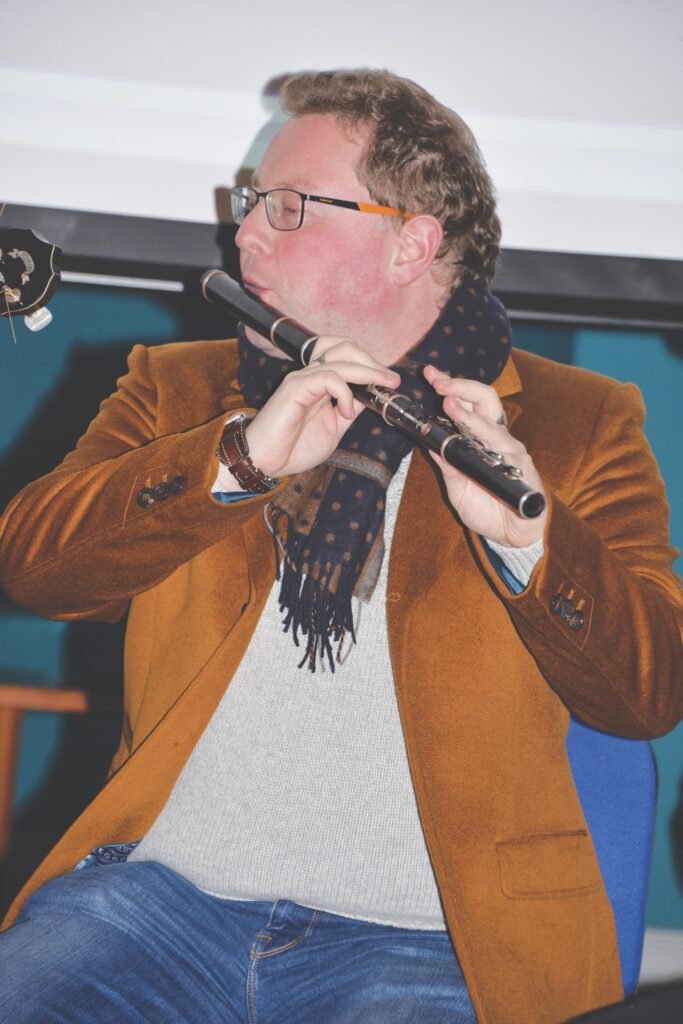 A seated man wearing glasses, a brown jacket and a scarf is playing the flute