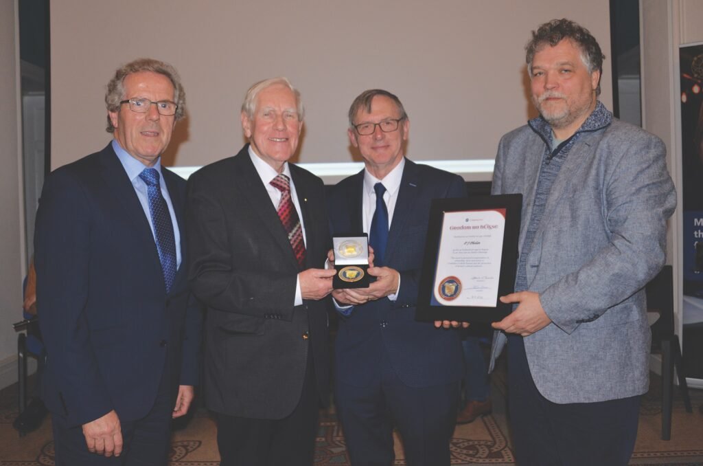 Four standing smiling men, three dressed in suits and ties, one wearing a suit jacket over a sweater and holding a framed certificate, the two men in the middle holding a medal between them