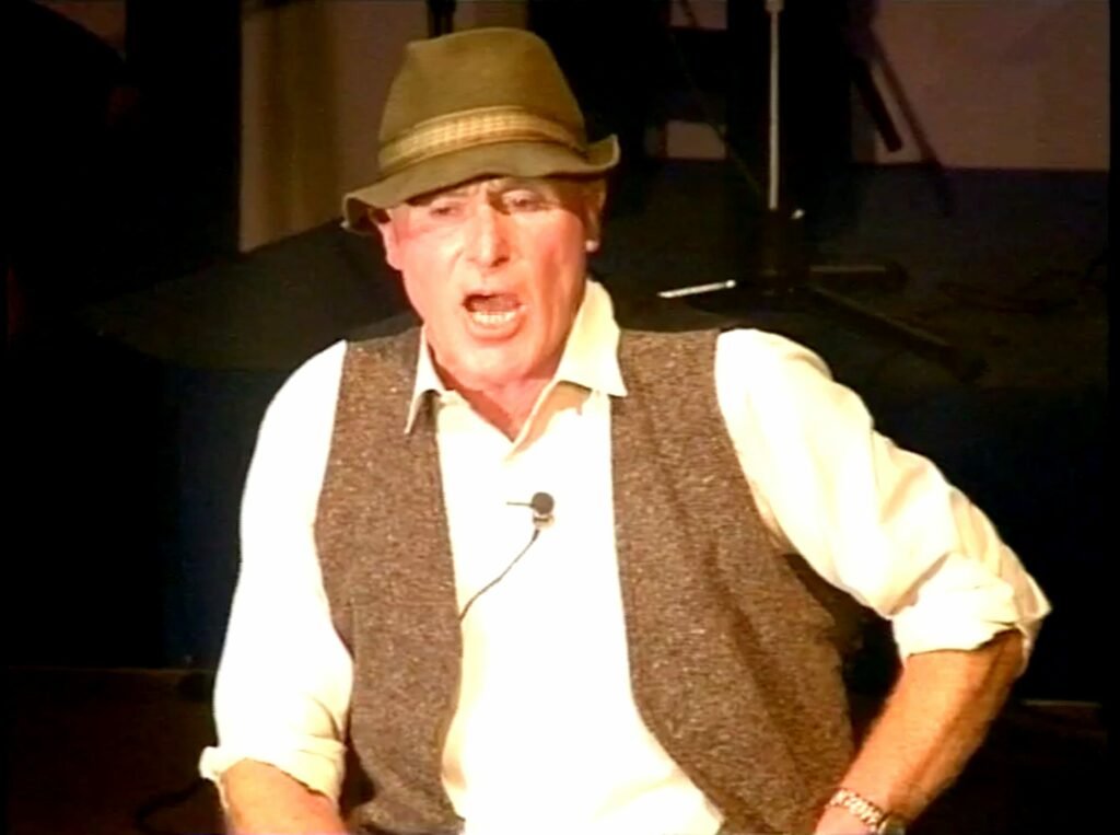 Colour photo of a man singing with a small microphone clipped to his white shirt, he is also wearing a brown waistcoat and hat