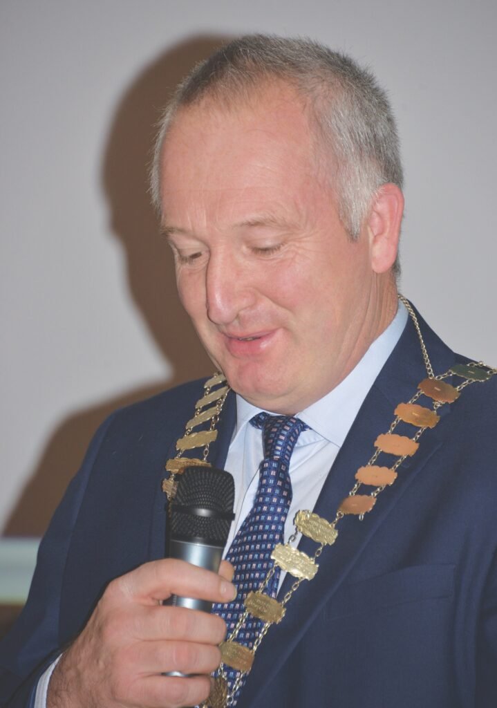 A man in a blue suit and tie looks down at a microphone he is holding, he is wearing a mayoral golden chain
