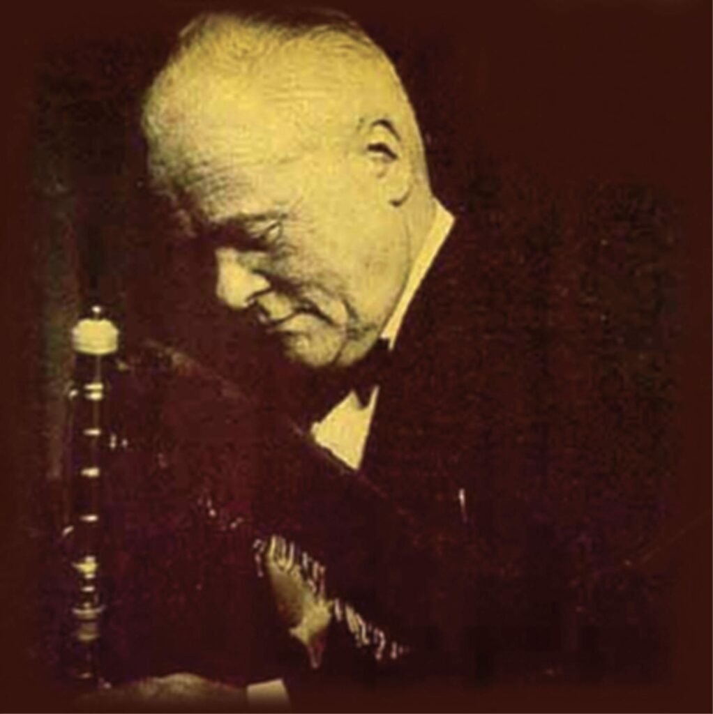 Sepia-toned photo of a man in side view looking down, the pipe of bagpipes is in front of him