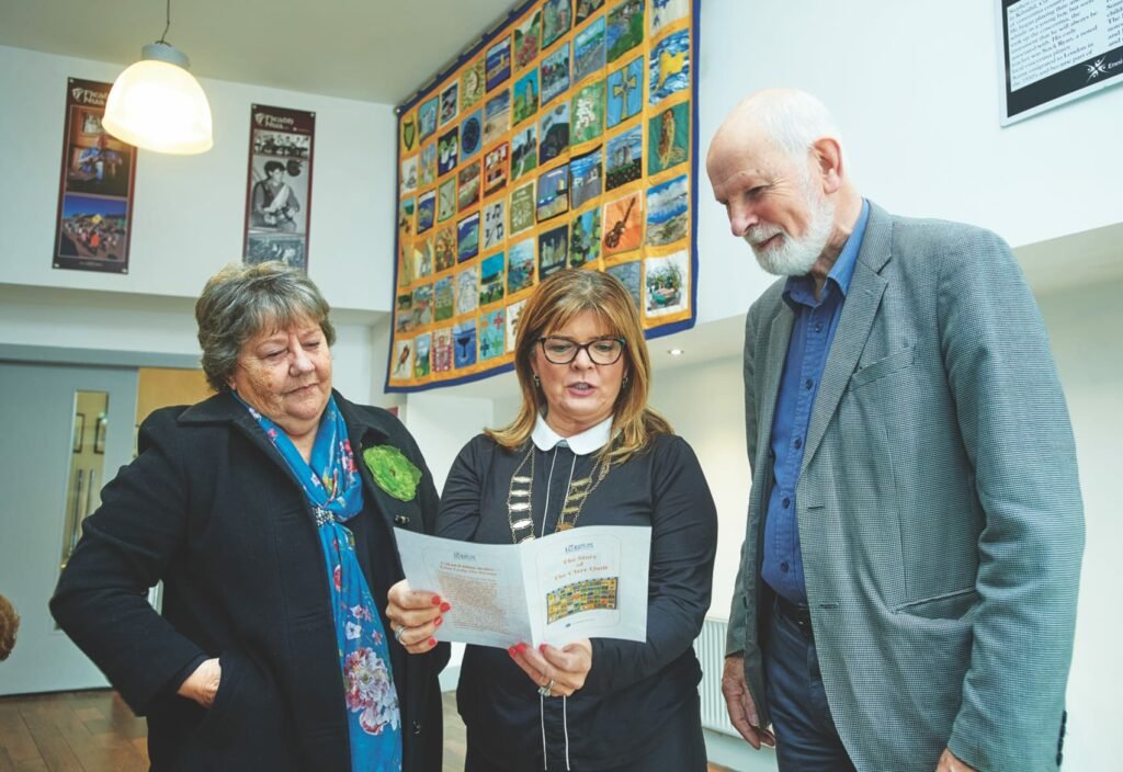 Two women and a man standing, looking at a booklet held by the woman in the middle who wears a mayoral chain of office, a large quilt with many panels hangs on the wall behind them