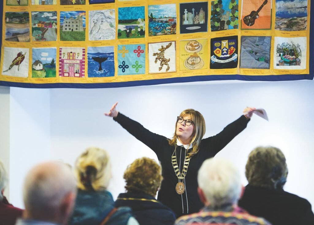 A woman weaing a mayoral chain of office, stands with her arms extended to each side, she is facing the camera, in the foreground are the backs of heads of the audience she is addressing, the bottom of a large quilt with many panels hangs on the wall behind her