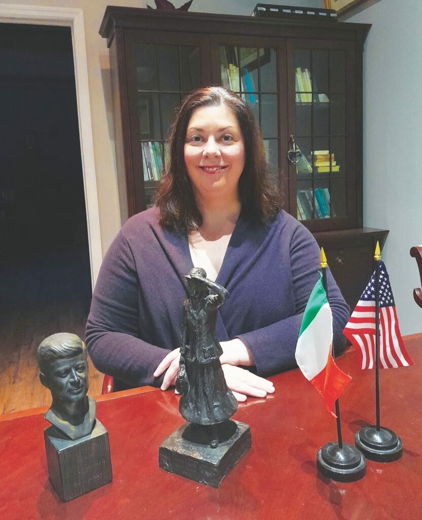 A smiling woman with long hair sits at a red desk, her hands crossed on the table, in front of her are small Irish and American flags, a small bust, and a small statuette of a woman