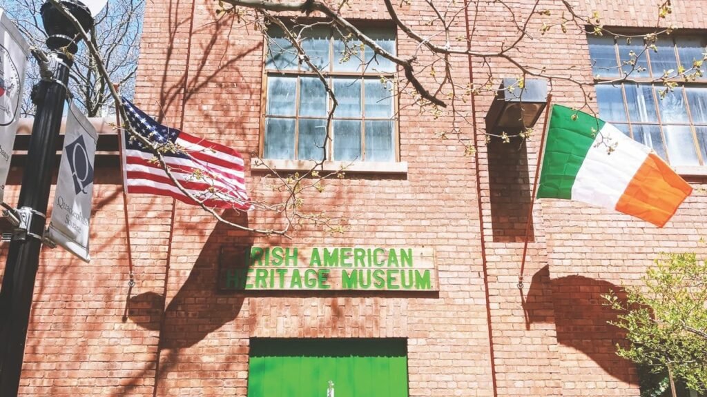 Part of the front of a building with Irish American Heritage Museum in green upper case lettering above the door, with an American flag to the left, an Irish flag to the right