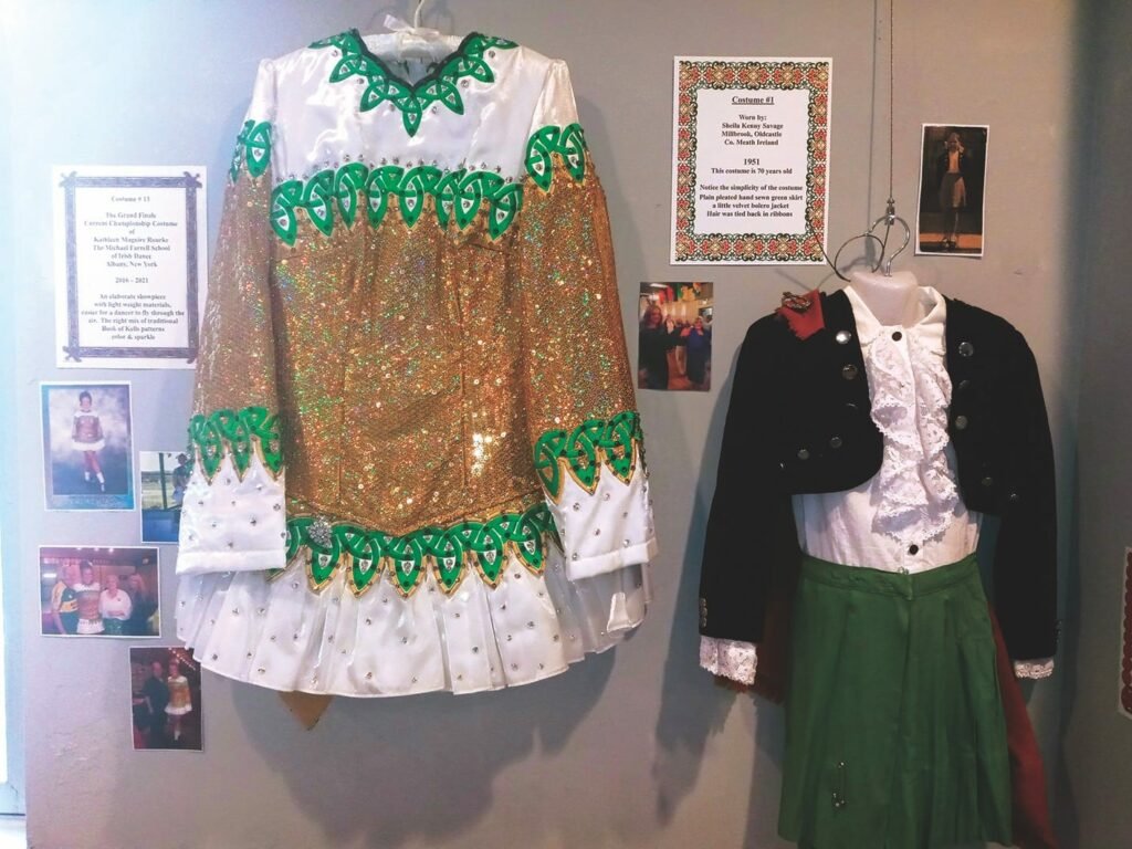 Two costumes hanging on display, the one on the left is a solid green kilt, a black jacket and a white shirt with a lace front and cuffs, the one on the left is a long-sleeved top with gold sequins across the middle, trimmed with white and green, and rhinestones, on the wall behind them are information panels and small photographs