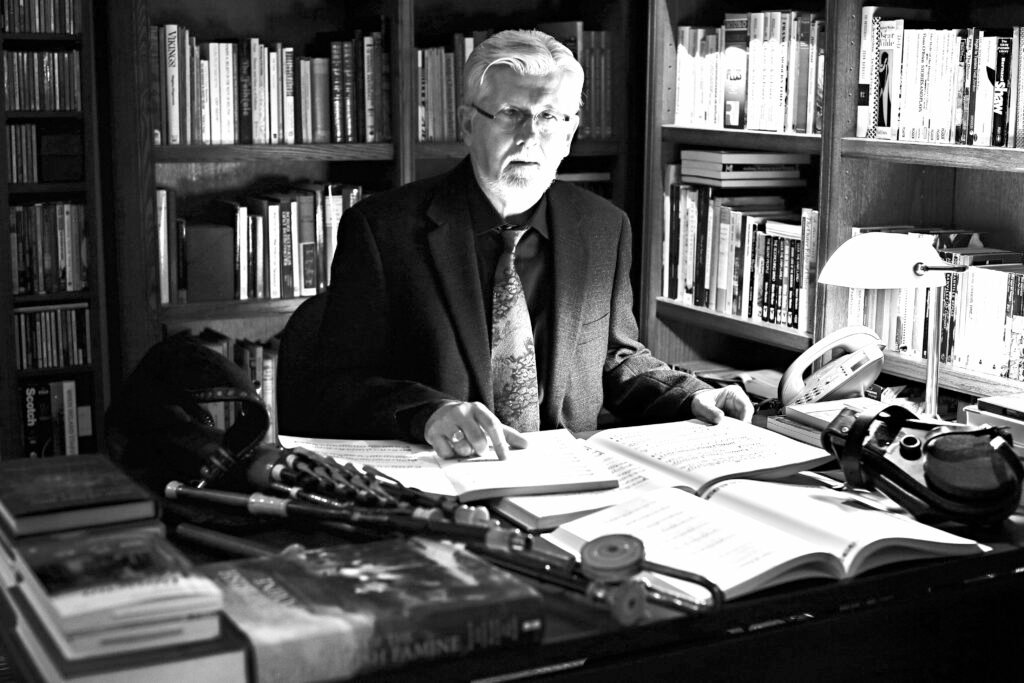 Black and white photo of a man with glasses, white hair and beard, in a book line room sitting with his hands on an open book on the desk in front of him, also on the desk are more books, some open, and bagpipes