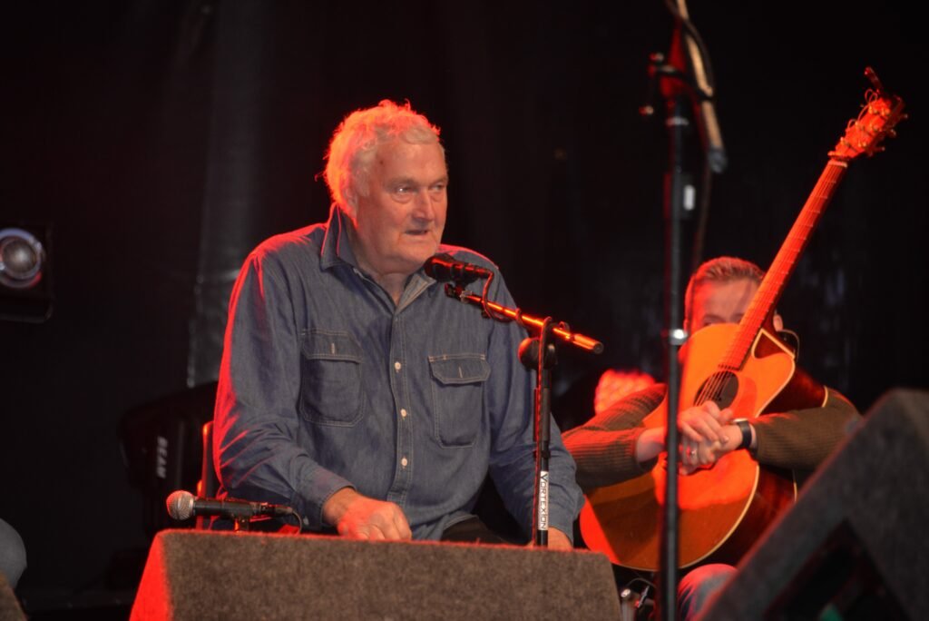 Colour photo of a man in a grey shirt singing into a microphone, to his left a man is sitting holding an acoustic guitar in front of his face