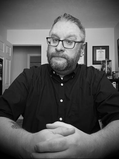 Author Bucho Rodenberger wearing a black button-up shirt sits at his desk in his home office with hands clasped together