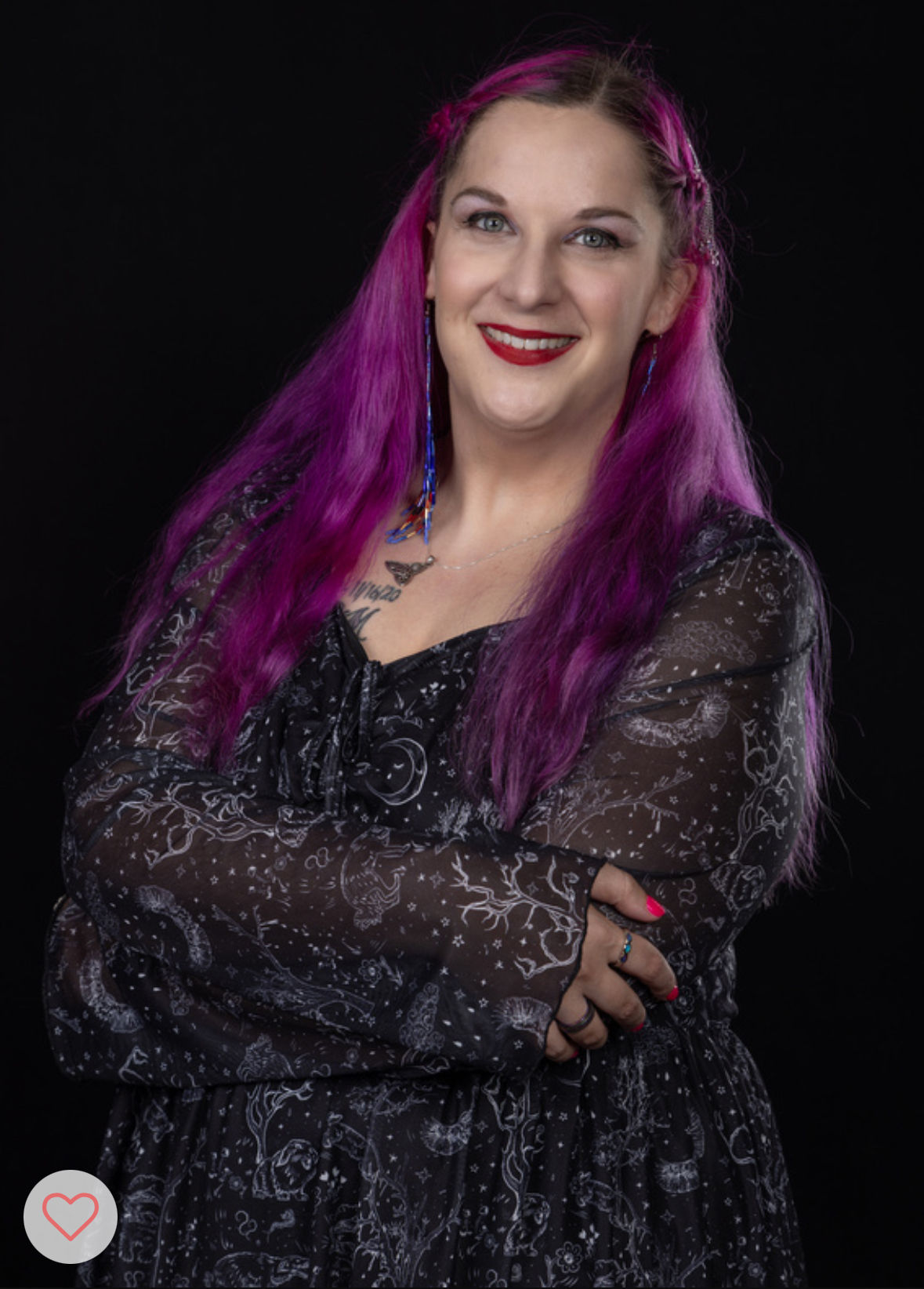 Woman in her 30s. Pink/purple wavy hair. Blue eyes with dark shadow and red lipstick. Dangling earrings. Wearing moon/stars black dress. Standing with arms crossed and smiling.