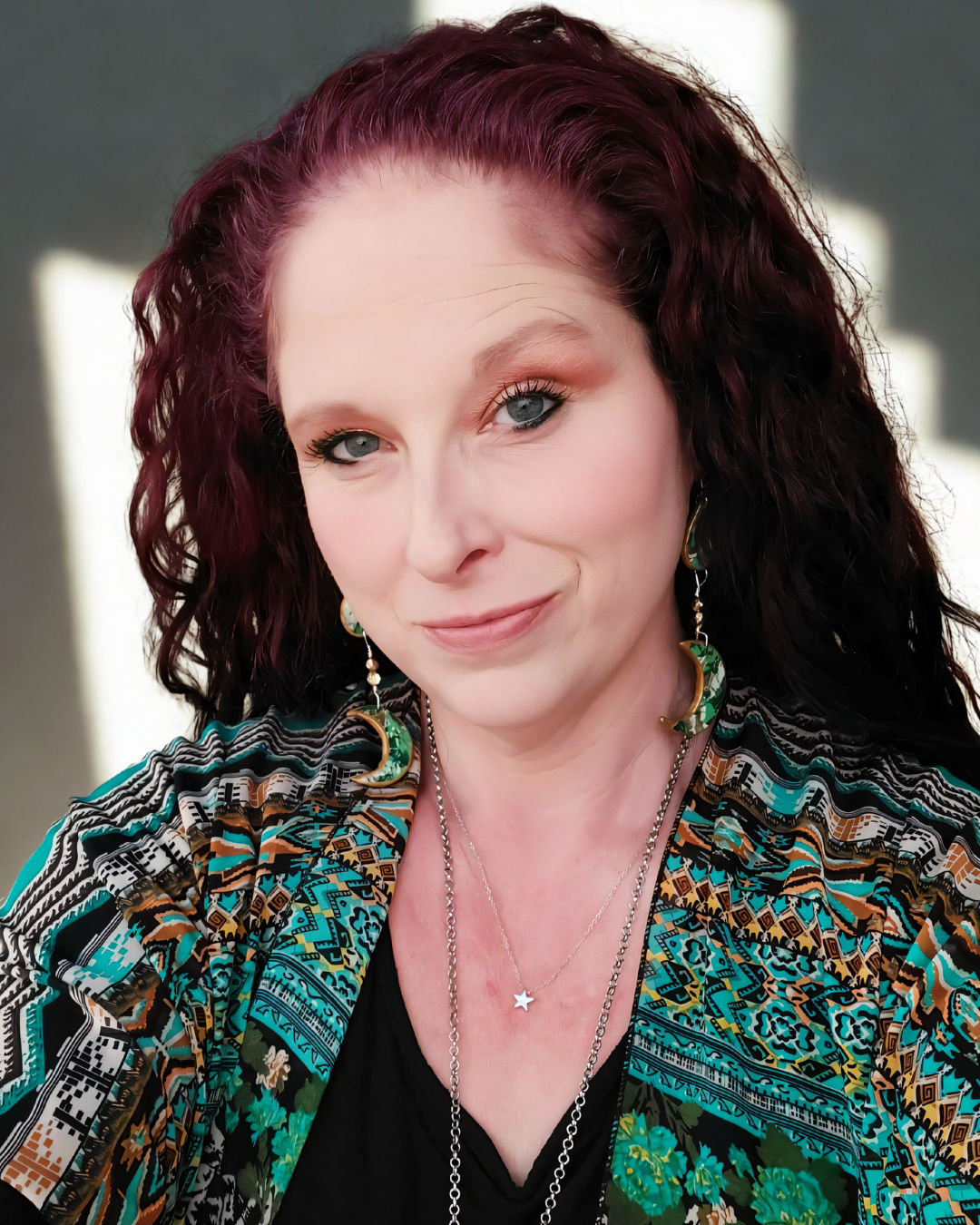 Alt text: Headshot of author MS Chambers, a woman with long, wavy auburn hair, wearing crescent moon earrings and a teal geometric print kimono.
Brief visual description: A close-up headshot of author MS Chambers looking forward with a slight smile. She has long, dark auburn, wavy hair and is wearing a black top paired with a vibrant, geometric-patterned kimono in teal, gold, and orange. Her accessories include long crescent moon earrings and a delicate star pendant necklace.