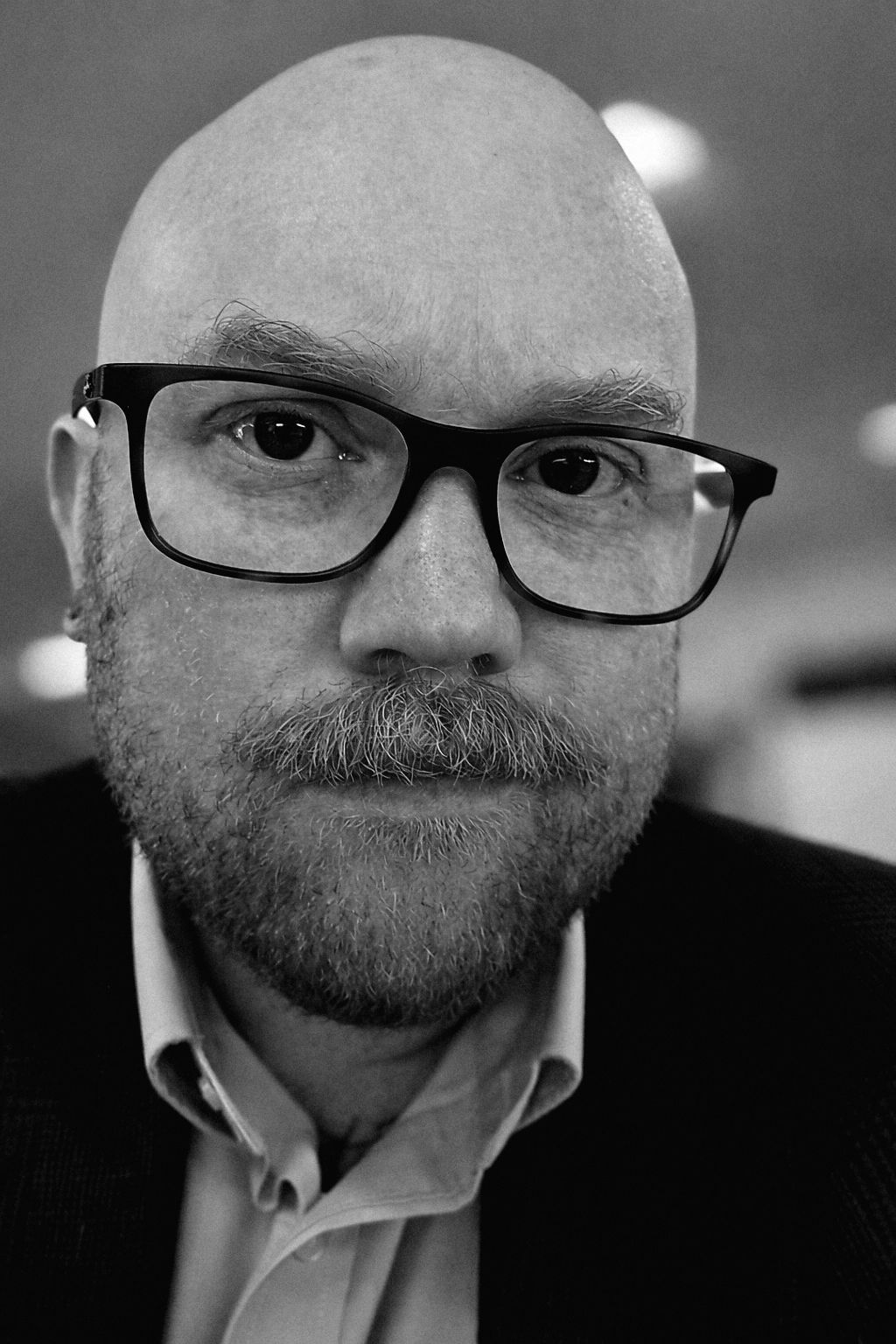 A bald, forty-something-year-old man whose eyesight is failing stares blankly at a camera lens, trying to look "authory".