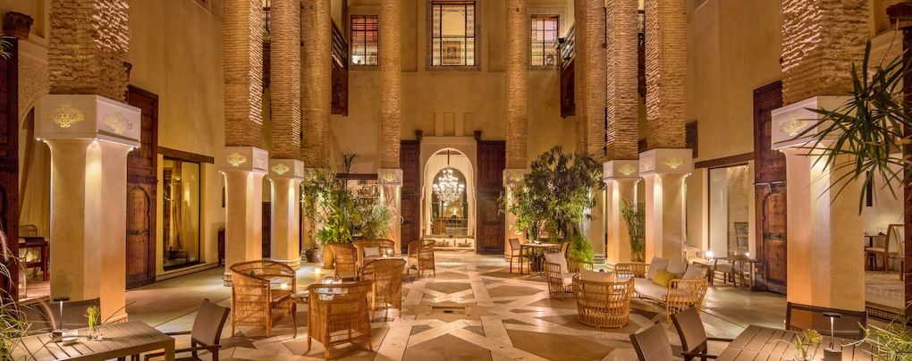 a well-lit and luxurious-looking courtyard with wooden chairs and tables in a high-class Moroccan riad