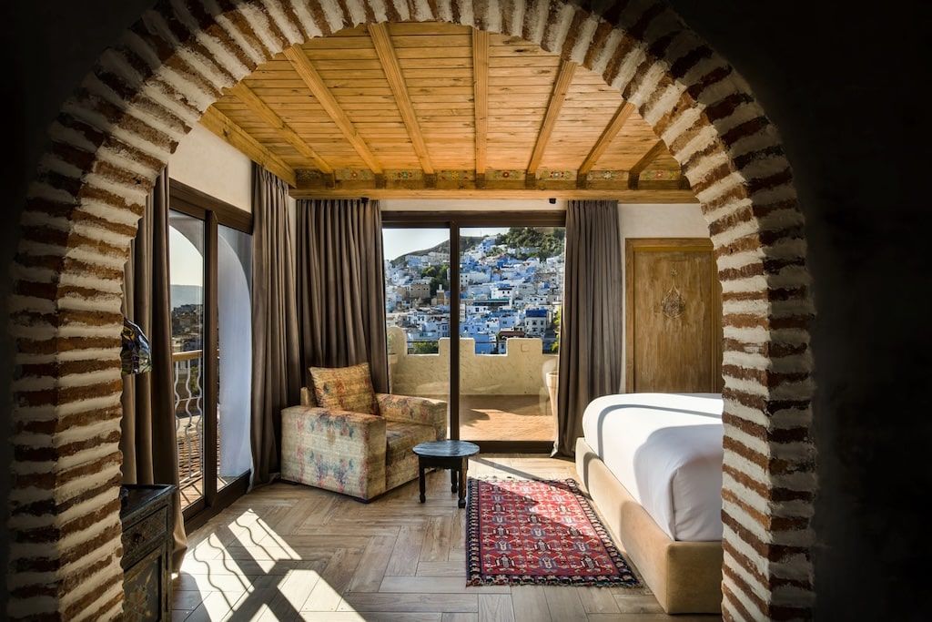 a framed view into a modernly-furnished room inside a riad with a view of a Moroccan Medina outside of the window in the background