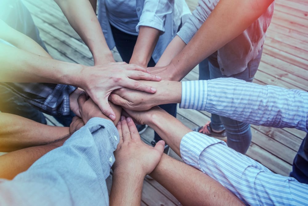 Hands from several people are all brought together stacked one on top of another as if a team is about to throw their hands up together in a cheer.