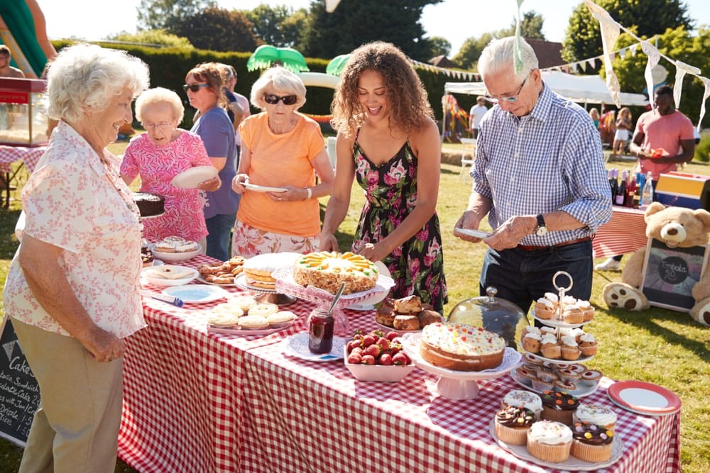 A group of smiling community members surround a table covered in baked goods to transact over purchases for a bake sale.