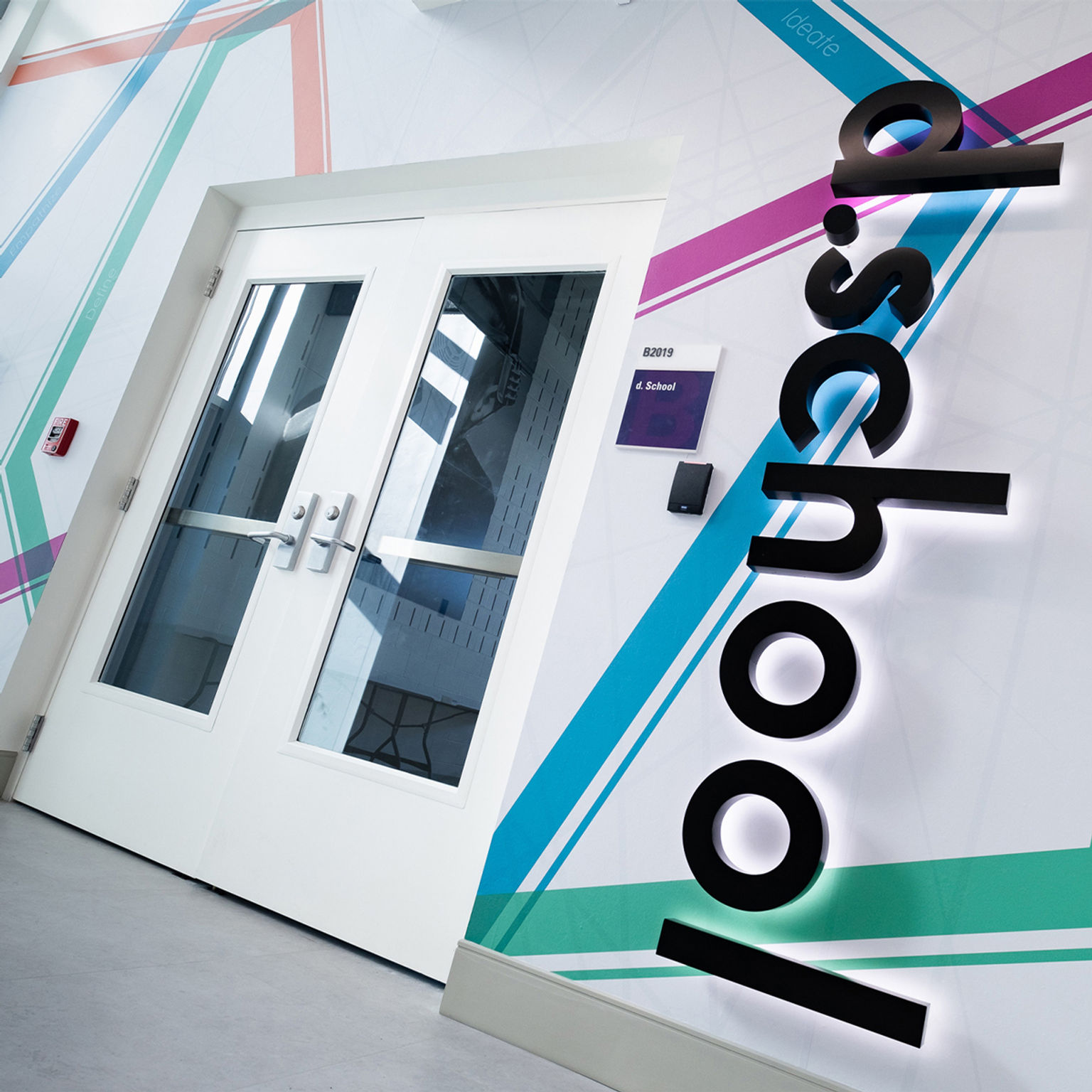 Low perspective view of experiential graphics installation surrounding the entrance of a design school entry featuring colorful custom wall covering, room identification signage, and illuminated vertically-oriented dimensional letting spelling "d.school."