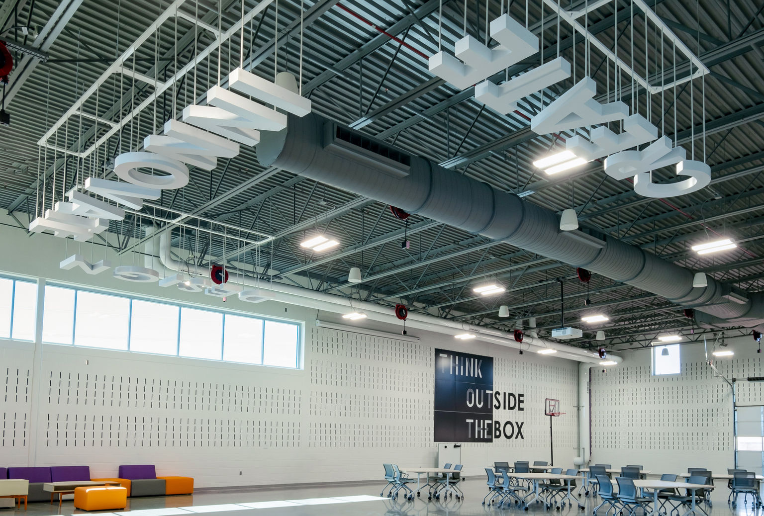 An expansive collaborative education space featuring white suspended dimensional letters spelling inspirational words such as "create," "design," and Innovate." Also featured is a dimensional wall graphic stating "Think outside the Box."