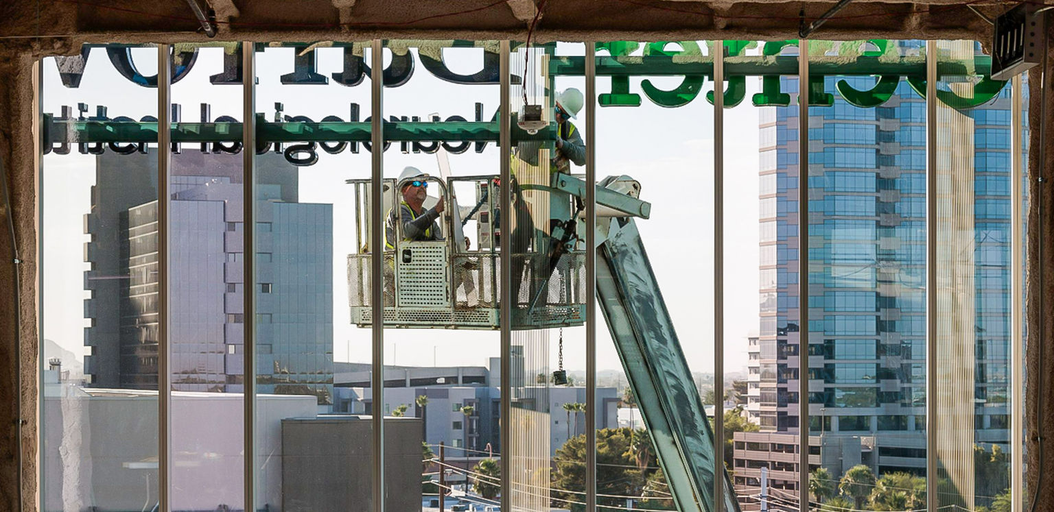 A dramatic inside-looking-out view of sign installers in a lift working on an on-building signage installation many floors above the ground.