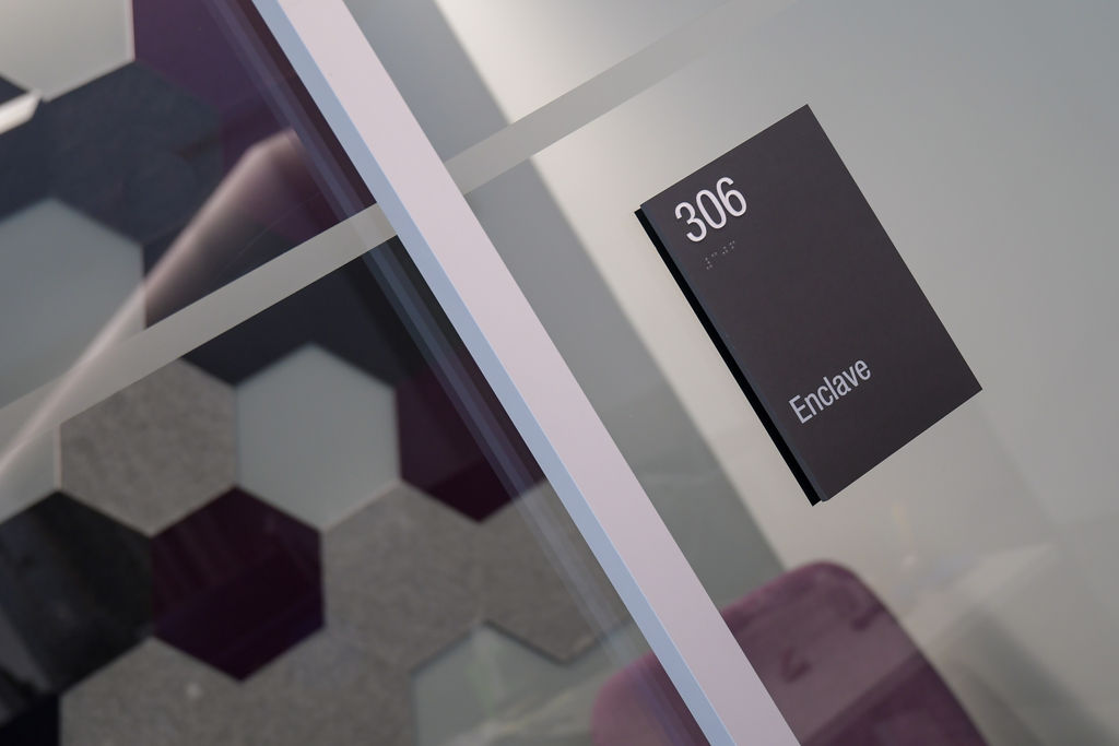 Interior glass-mounted ADA compliant Room ID signage with colorful acoustic tiles visible in the background.