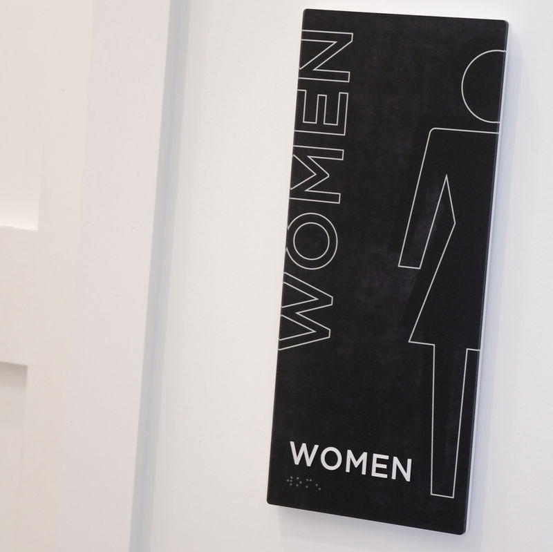 A black and white women's room sign with a contemporary minimalist design made with Vivid construction is easy to clean because it has no edges or crevices.