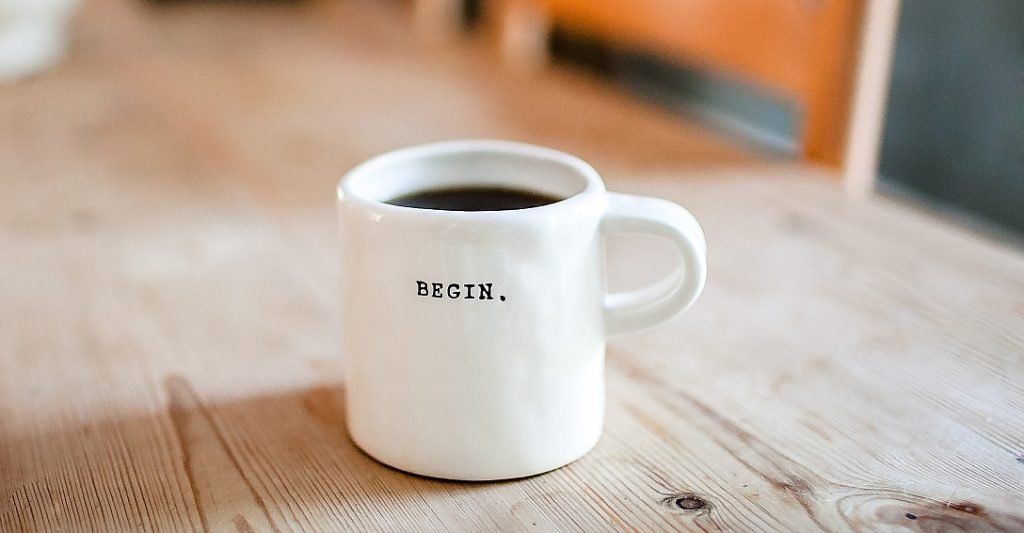 A cup of coffee with the word 'Begin' printed on it placed on a wooden table. Photo by Danielle MacInnes on Unsplash