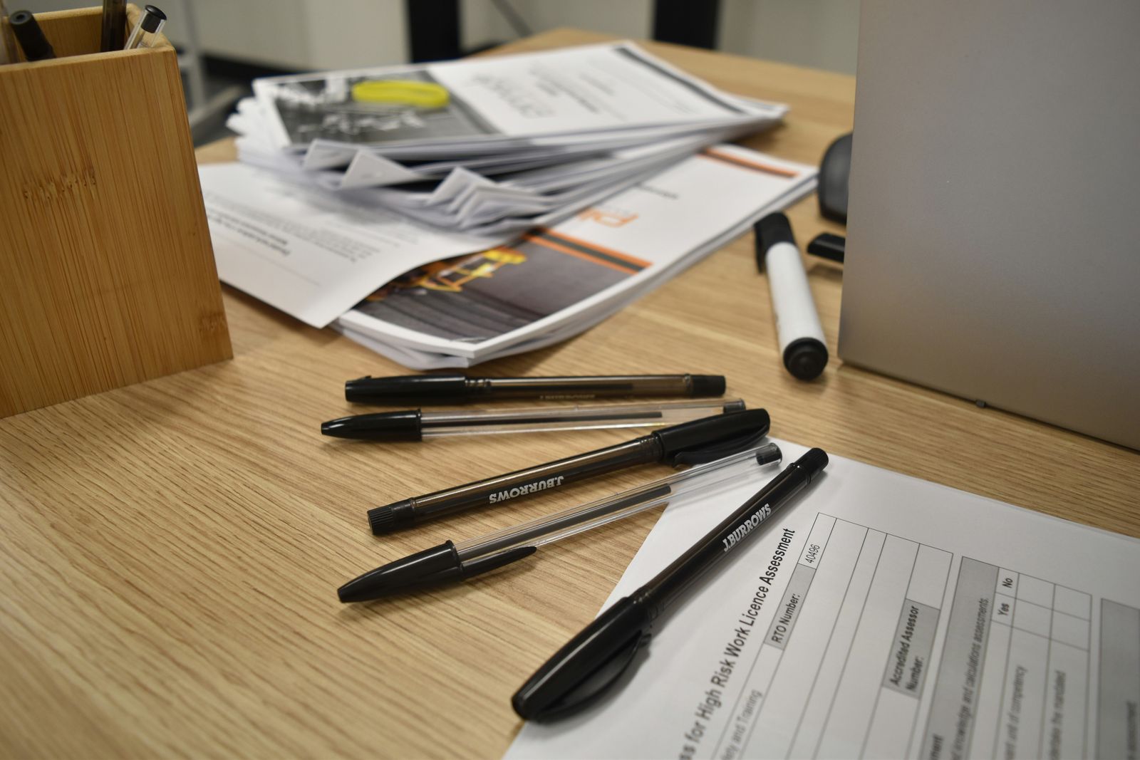 Close-up of paperwork and pens on a wooden desk next to an open laptop